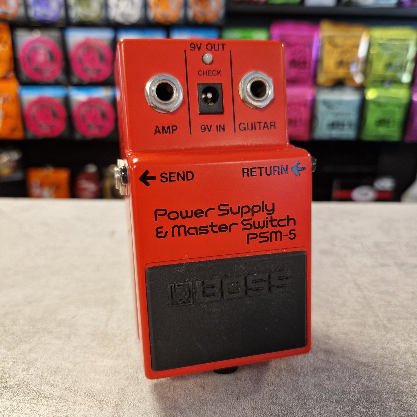 Pre-Owned Boss PSM-5 Red Boss guitar pedal on a textured surface with colorful items in the background