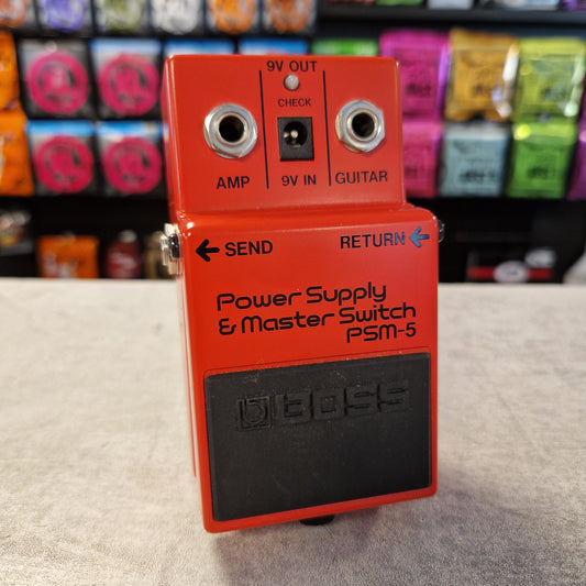 Pre-Owned Boss PSM-5 Red Boss guitar pedal on a textured surface with colorful items in the background
