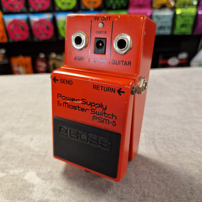 Pre-Owned Boss PSM-5 Red Boss power supply switch on a textured surface with colorful boxes in the background