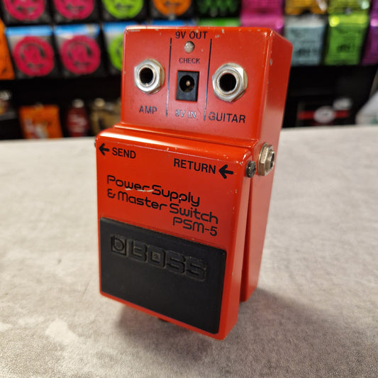 Pre-Owned Boss PSM-5 Red Boss power supply switch on a textured surface with colorful boxes in the background