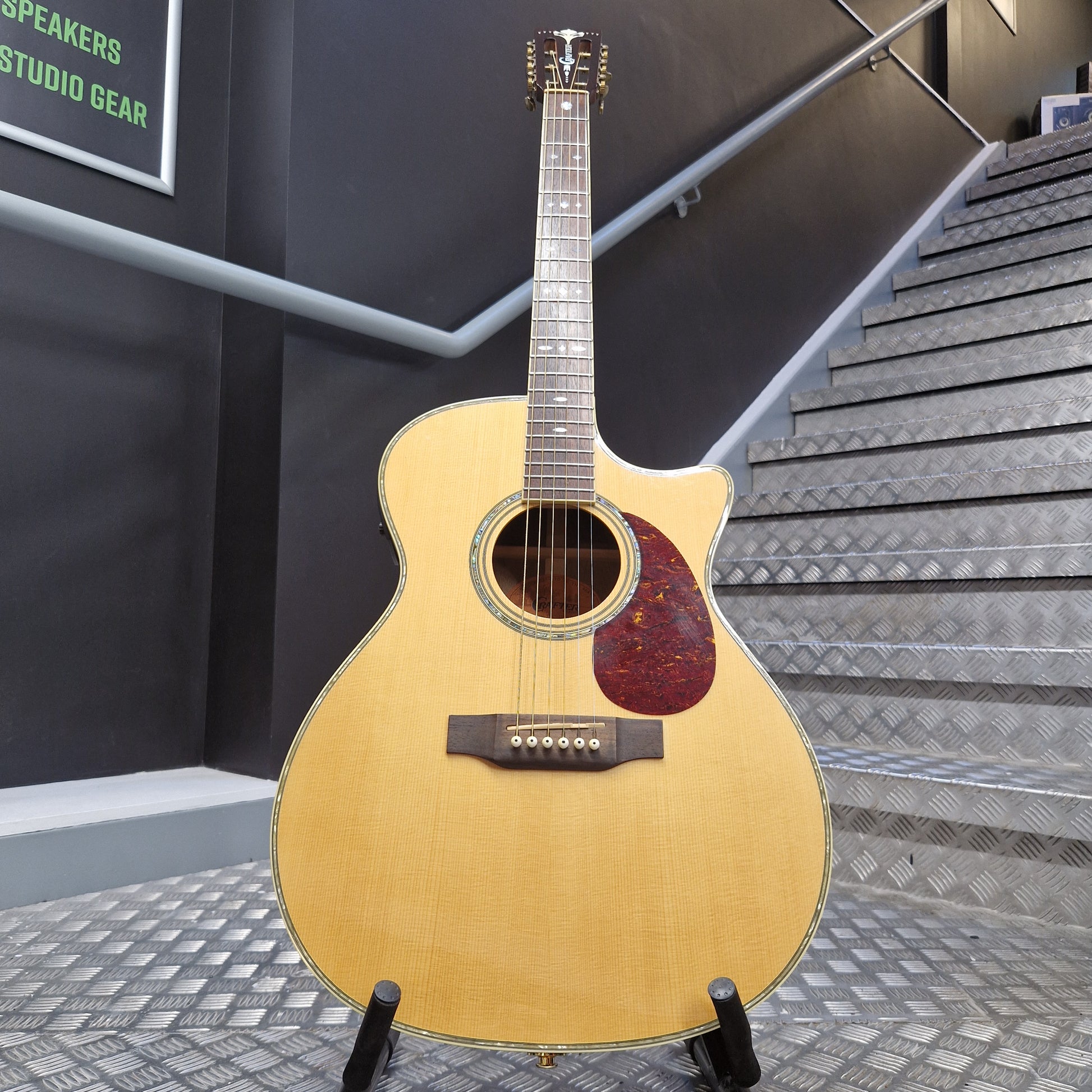 Pre-Owned Crafter TMC035/N Acoustic guitar on a stand in front of a staircase with a dark wall and metal railing.
