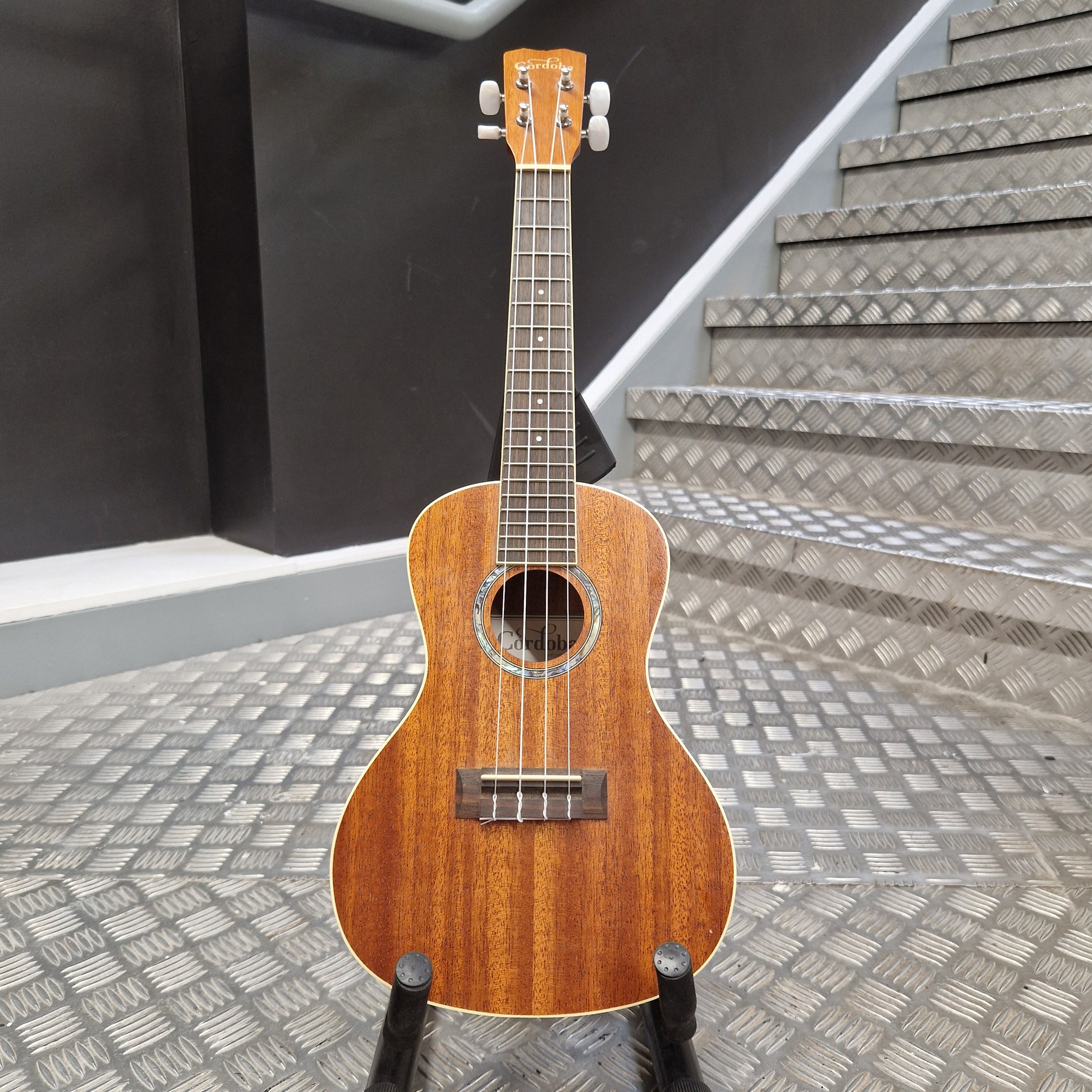 Pre-Owned Cordoba 15CM Wooden ukulele on a stand with a staircase in the background