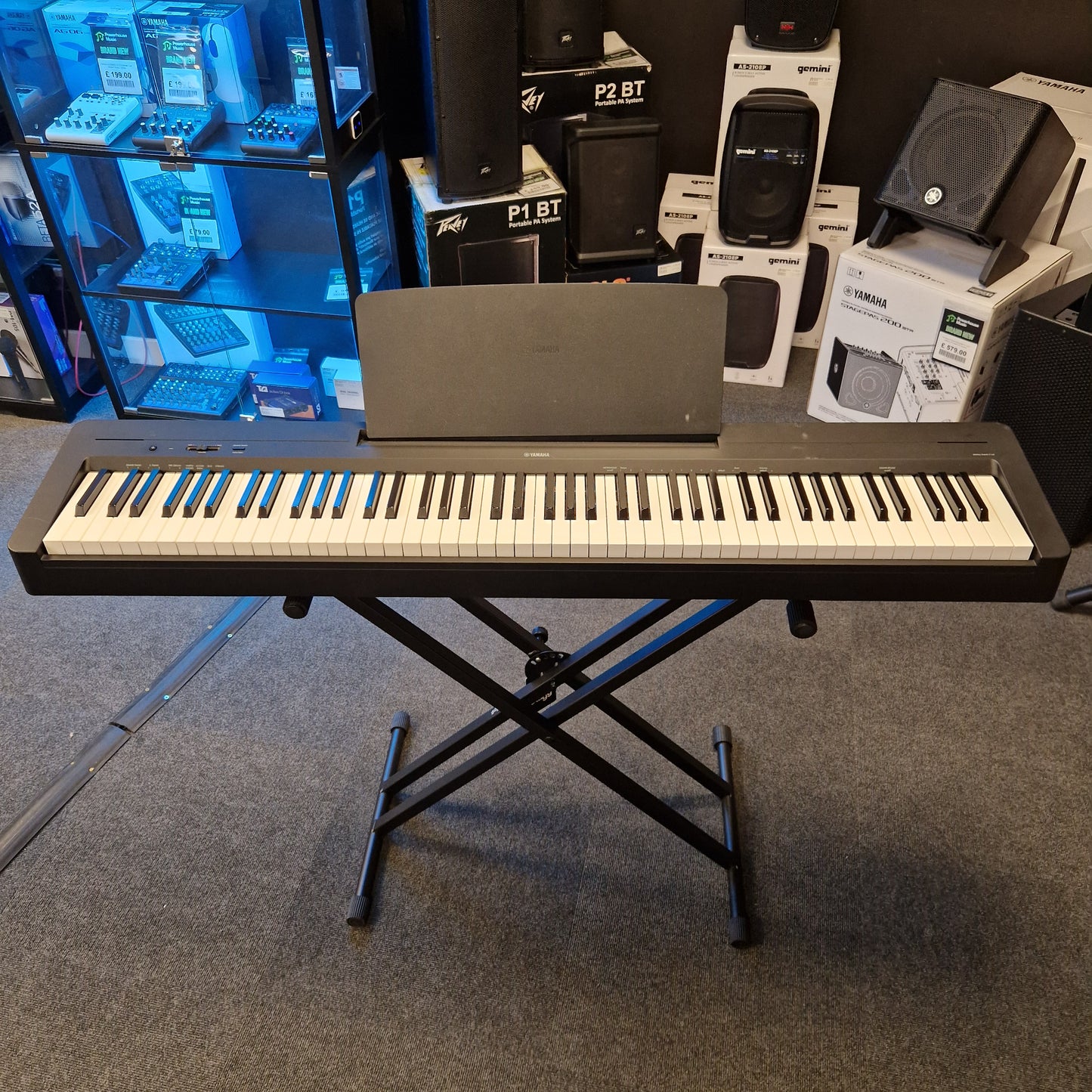 Pre-Owned Yamaha P-145B Keyboard on a stand in a room with electronic equipment and shelves.