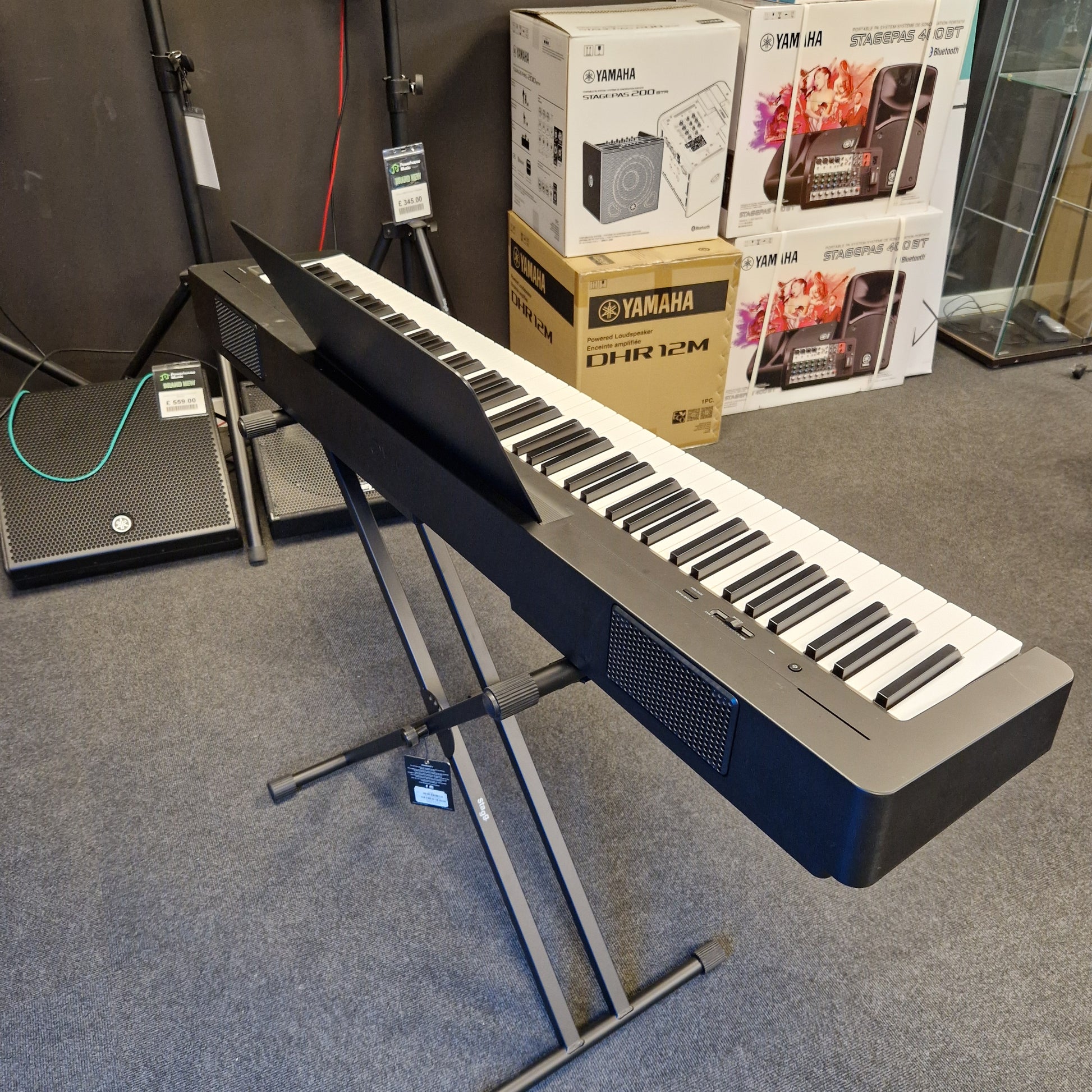 Pre-Owned Yamaha P-145B Yamaha keyboard on a stand with boxes and magazines in the background