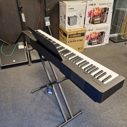 Pre-Owned Yamaha P-145B Yamaha keyboard on a stand with boxes and magazines in the background