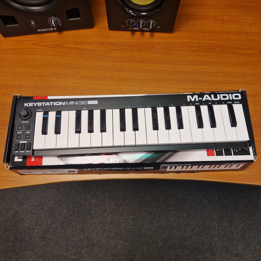 Pre-Owned M-Audio Keystation Mini 32 M-Audio keyboard on a wooden surface with audio equipment in the background