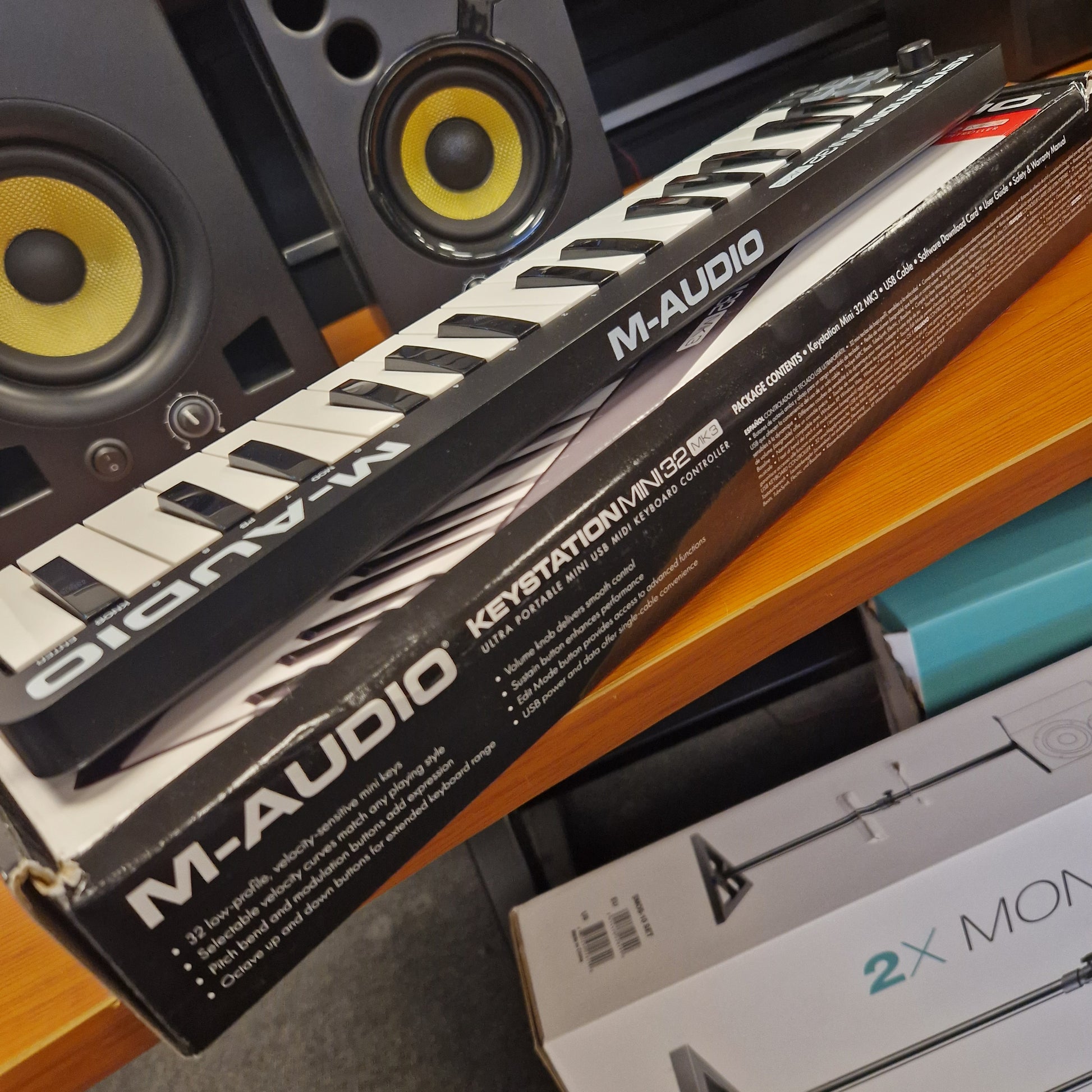Pre-Owned M-Audio Keystation Mini 32 M-Audio keyboard controller packaging on a surface with speakers and other equipment.