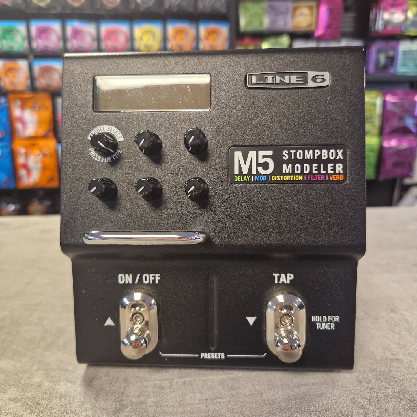 Pre-Owned Line 6 M5 Line 6 Stompbox Modeler M5 guitar effects pedal on a gray surface with colorful boxes in the background.