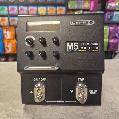 Pre-Owned Line 6 M5 Line 6 Stompbox Modeler M5 guitar effects pedal on a gray surface with colorful boxes in the background.