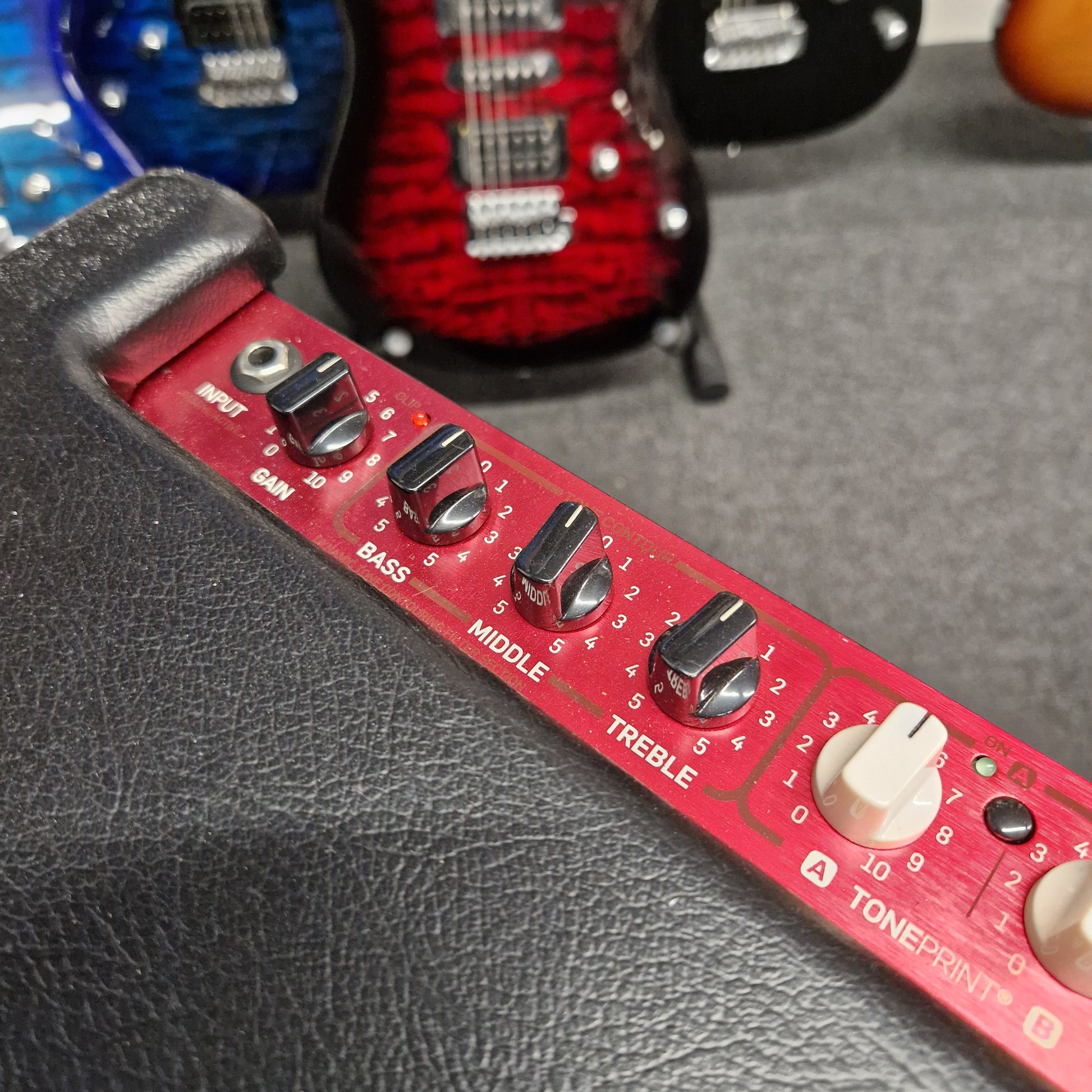 Pre-Owned TC Electronic BG250-112 Red guitar equalizer with knobs on a gray surface, with a red and black guitar in the background.