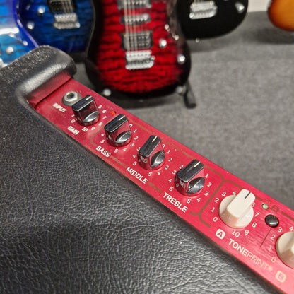 Pre-Owned TC Electronic BG250-112 Red guitar equalizer with knobs on a gray surface, with a red and black guitar in the background.