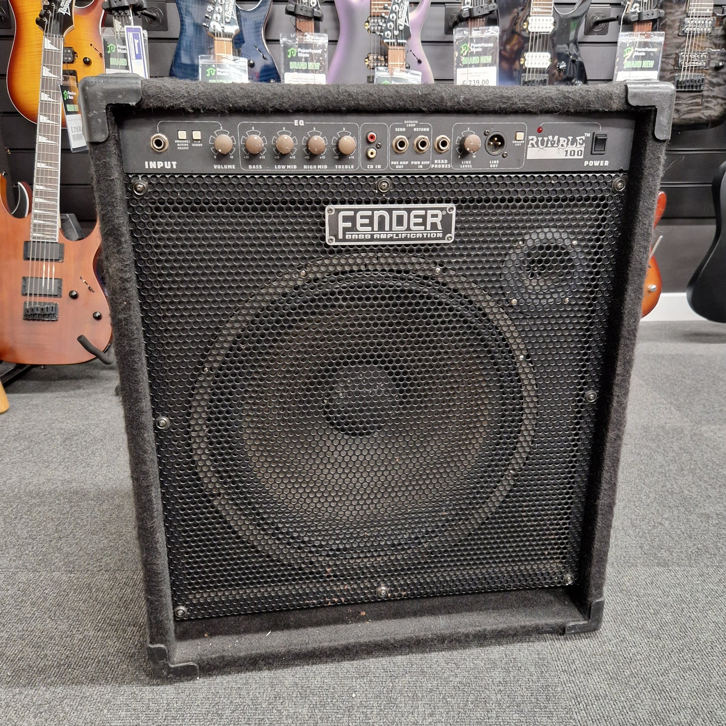 Pre-Owned Fender Rumble 100 Fender guitar amplifier on a gray surface with guitars in the background