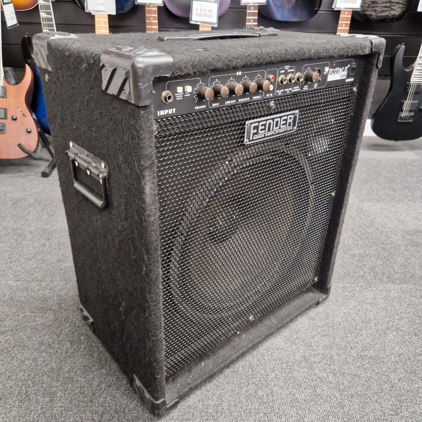 Pre-Owned Fender Rumble 100 Fender guitar amplifier on a gray floor with guitars in the background