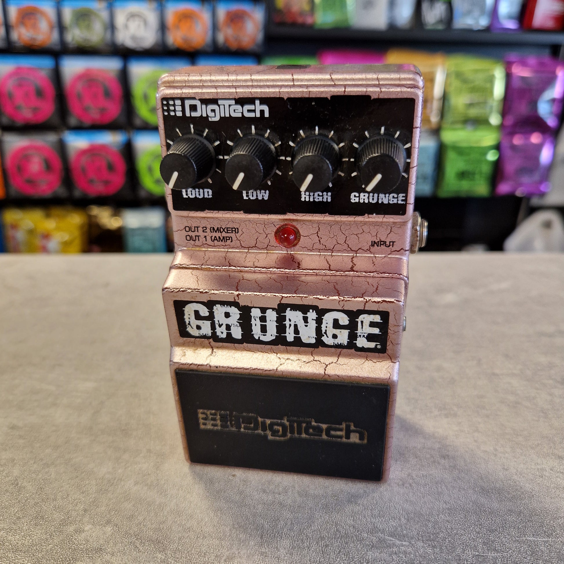 Pre-Owned Digitech Grunge A Digitech Grunge guitar effects pedal with a metallic rose gold/champagne body, black knobs, and a black section on the right side, photographed standing on a gray floor with colorful objects blurred in the background.