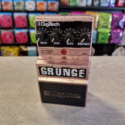 Pre-Owned Digitech Grunge A Digitech Grunge guitar effects pedal with a metallic rose gold/champagne body, black knobs, and a black section on the right side, photographed standing on a gray floor with colorful objects blurred in the background.
