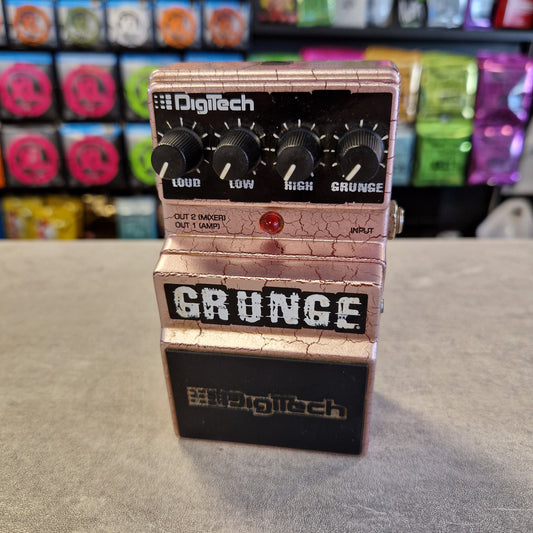 Pre-Owned Digitech Grunge A Digitech Grunge guitar effects pedal with a metallic rose gold/champagne body, black knobs, and a black section on the right side, photographed standing on a gray floor with colorful objects blurred in the background.