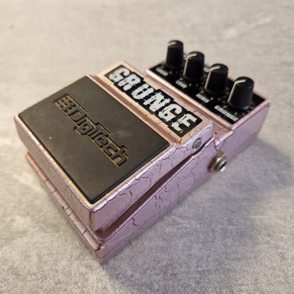 Pre-Owned Digitech Grunge A Digtech Grounge guitar effects pedal with a textured light purple/pink casing, black knobs, and a black footswitch plate, shown on a gray textured surface.