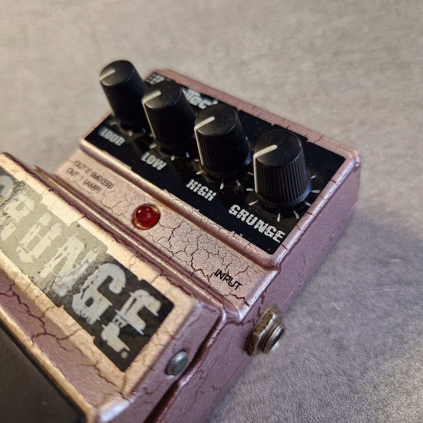 Pre-Owned Digitech Grunge A close-up angled shot of a guitar effects pedal with a cracked rose-pink metal casing, black knobs, a red indicator light, and a vintage-style label.