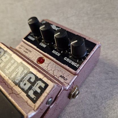 Pre-Owned Digitech Grunge A close-up angled shot of a guitar effects pedal with a cracked rose-pink metal casing, black knobs, a red indicator light, and a vintage-style label.