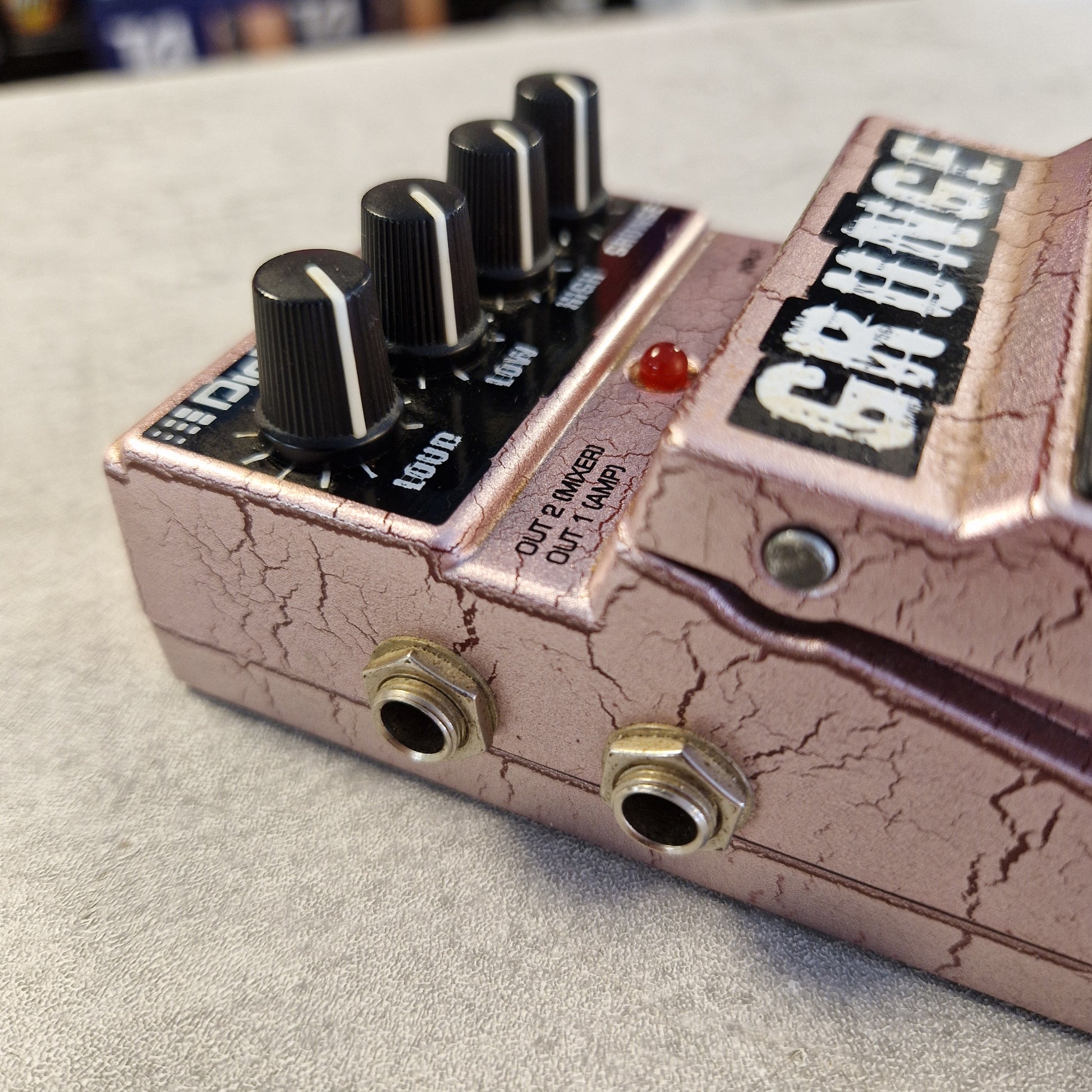 Pre-Owned Digitech Grunge Close-up of a guitar effects pedal with a cracked metallic pinkish-bronze finish, black control knobs, and gold-toned jacks on a light textured surface.