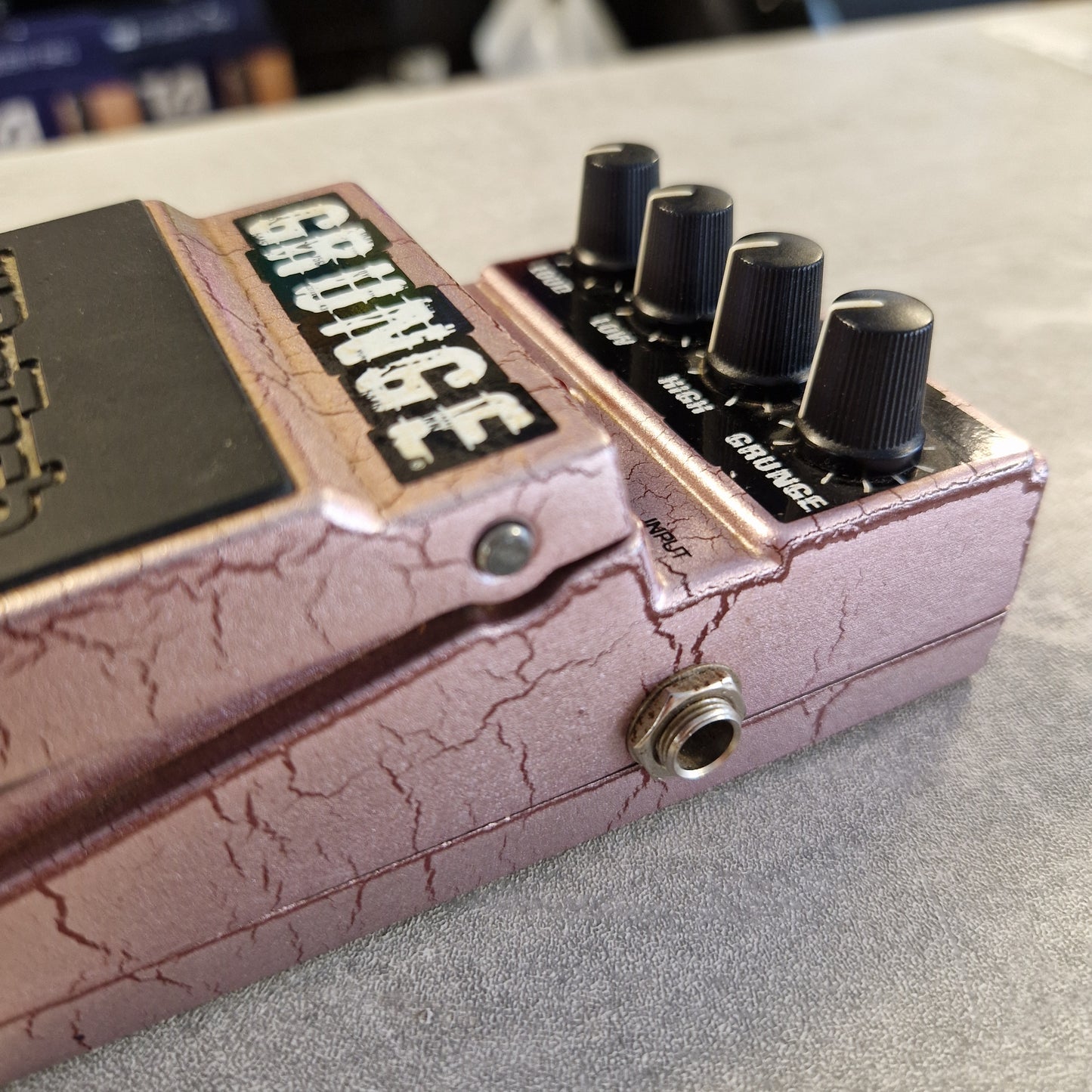 Pre-Owned Digitech Grunge A close-up of a guitar effects pedal labeled 'GRUNGE' with a cracked mauve-pink finish, black knobs, and a metal input jack, photographed against a gray textured surface.