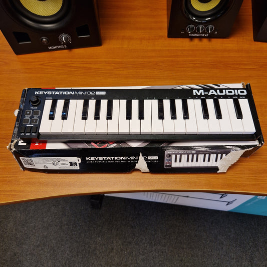 Pre-Owned M-Audio Keystation A Korg Kronos Station 16 MIDI controller in its original retail box, displayed on a wooden desk next to audio speakers.
