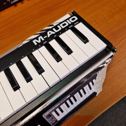 Pre-Owned M-Audio Keystation An M-Audio MKS-1000 midi controller with black and white keys, shown partially wrapped in protective plastic on a wooden table.