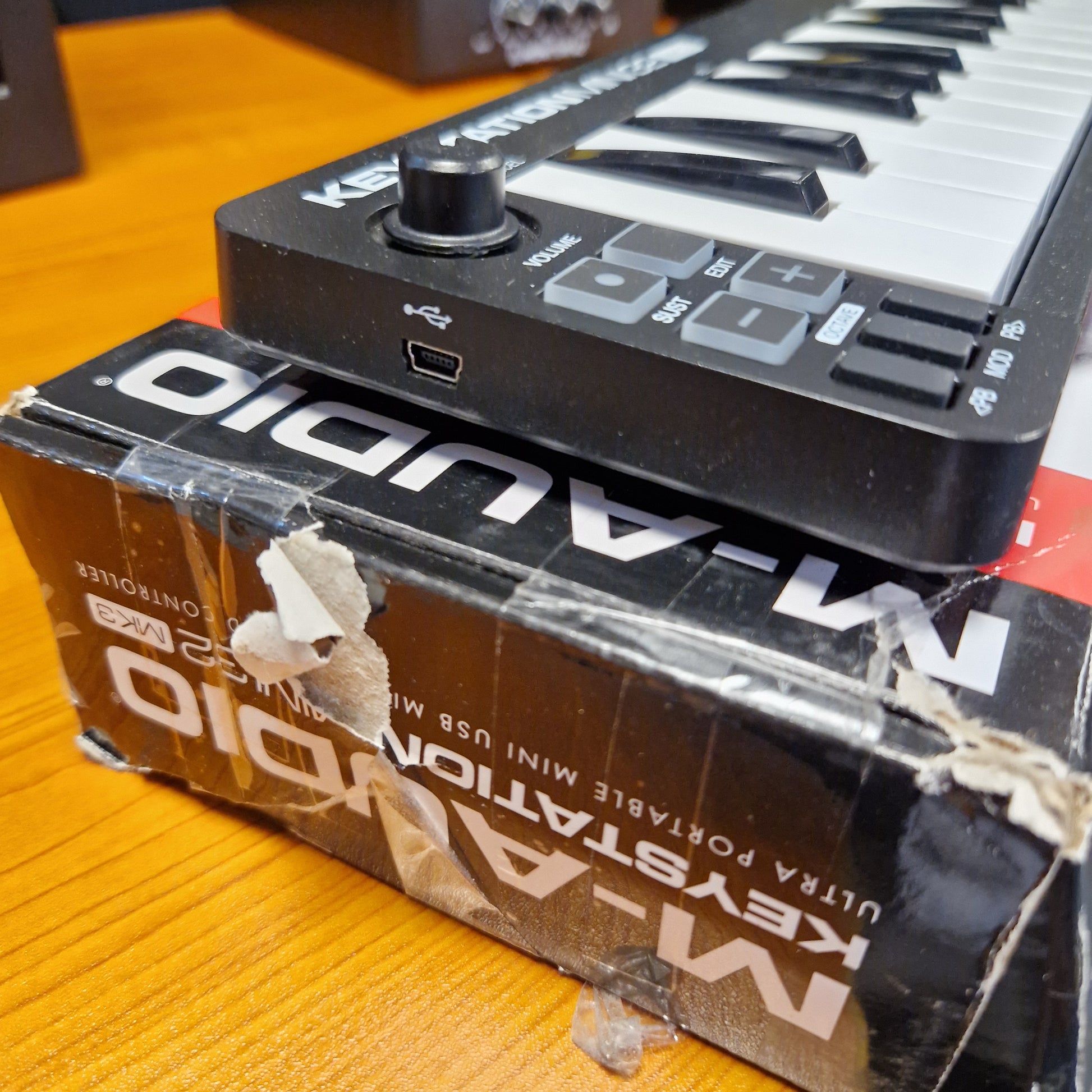 Pre-Owned M-Audio Keystation A close-up of a small black and white mini keyboard resting on top of its original retail box, which is placed on a wooden surface.