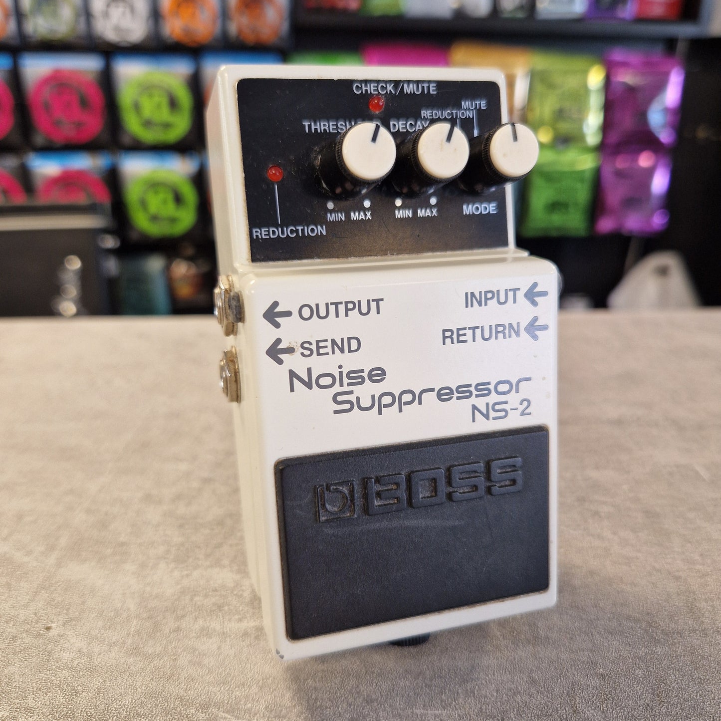 Pre-Owned Boss NS-2  A Boss NS-2 Noise Suppresser guitar pedal with a cream-colored plastic body, black control panel, and white knobs, photographed on a textured gray surface with a blurred background of colorful effects pedals.