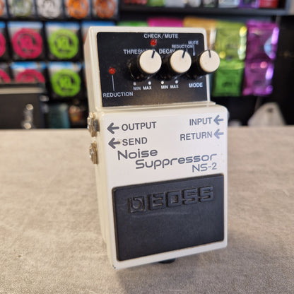 Pre-Owned Boss NS-2  A Boss NS-2 Noise Suppresser guitar pedal with a cream-colored plastic body, black control panel, and white knobs, photographed on a textured gray surface with a blurred background of colorful effects pedals.
