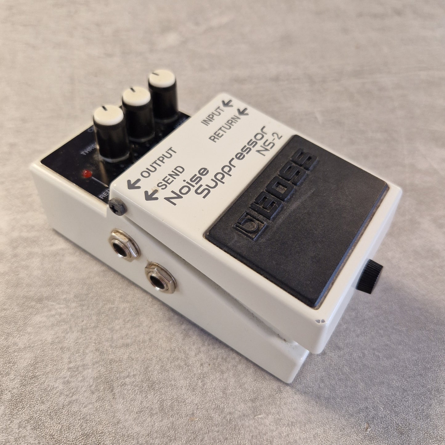 Pre-Owned Boss NS-2  A Boss NS-2 Noise Suppressor guitar pedal, shown at an angle on a textured gray surface. The pedal has a white plastic casing, a black footplate, three black volume knobs with white tops, and two gold-toned jack inputs.