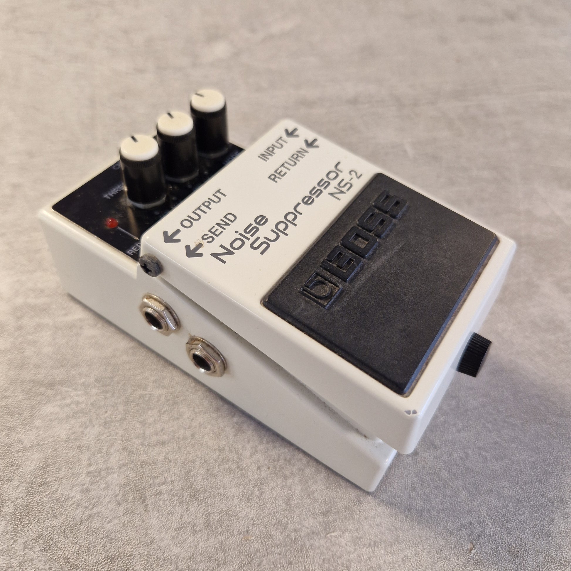 Pre-Owned Boss NS-2  A Boss NS-2 Noise Suppressor guitar pedal, shown at an angle on a textured gray surface. The pedal has a white plastic casing, a black footplate, three black volume knobs with white tops, and two gold-toned jack inputs.