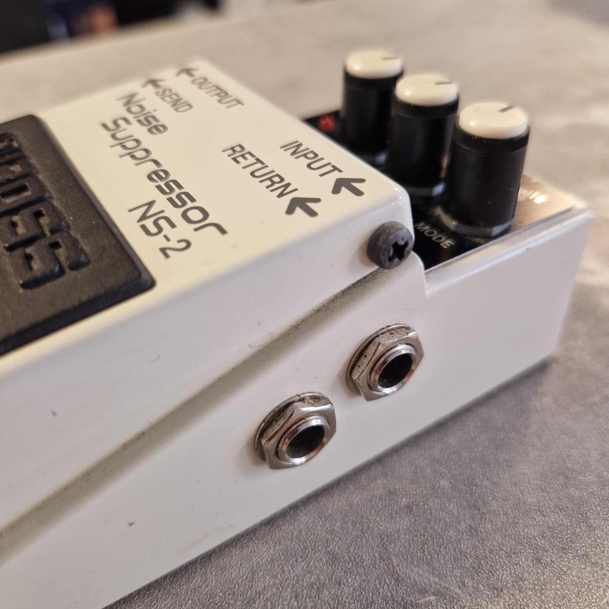 Pre-Owned Boss NS-2  Close-up of a white Boss noise suppressor pedal, showing the white casing, black knobs, silver jacks, and the Boss logo on a gray textured surface.