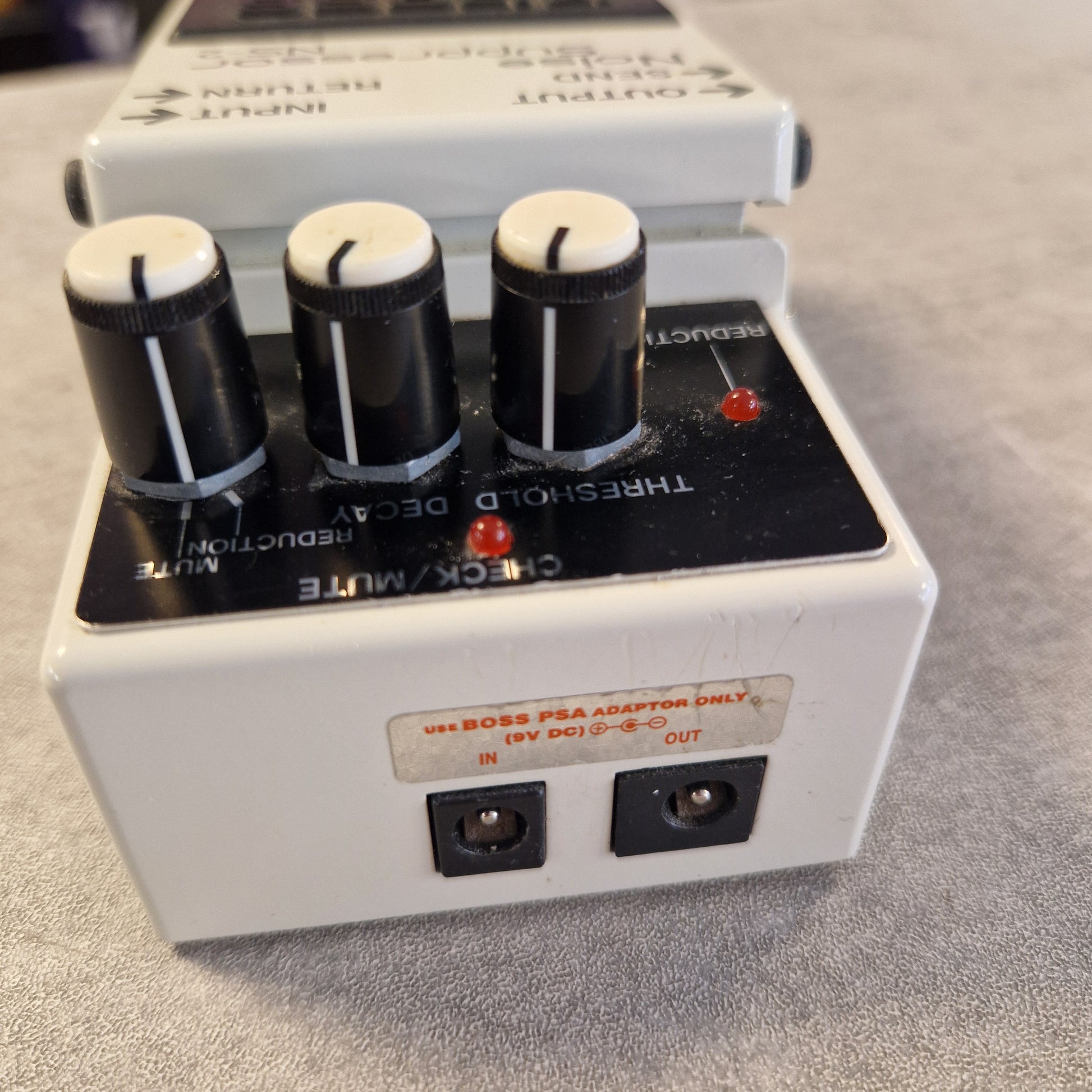 Pre-Owned Boss NS-2  A close-up of a white and black guitar effects pedal, showing three black knobs with white tops, red indicator lights, and a power jack on the side, photographed on a gray textured surface.