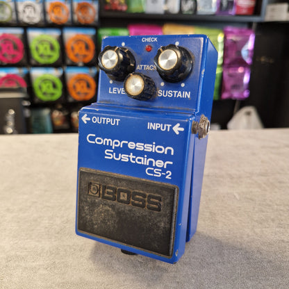 Pre-Owned Boss CS-2 A blue BOSS Compression Sustainer CS-2 guitar pedal with black knobs and a black footswitch panel, photographed on a light gray surface with a blurred background of colorful items.