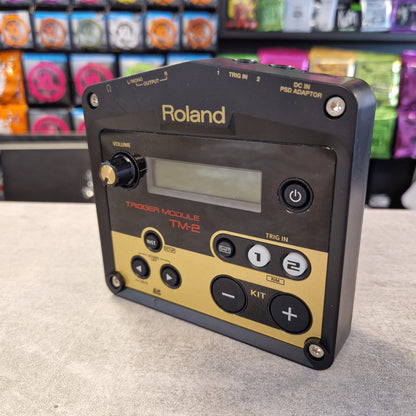 Pre-Owned Roland TM-2 A Roland TD-1000 Time Module electronic device with a black casing and a gold control panel, photographed on a light gray surface with a blurred background of colorful items.
