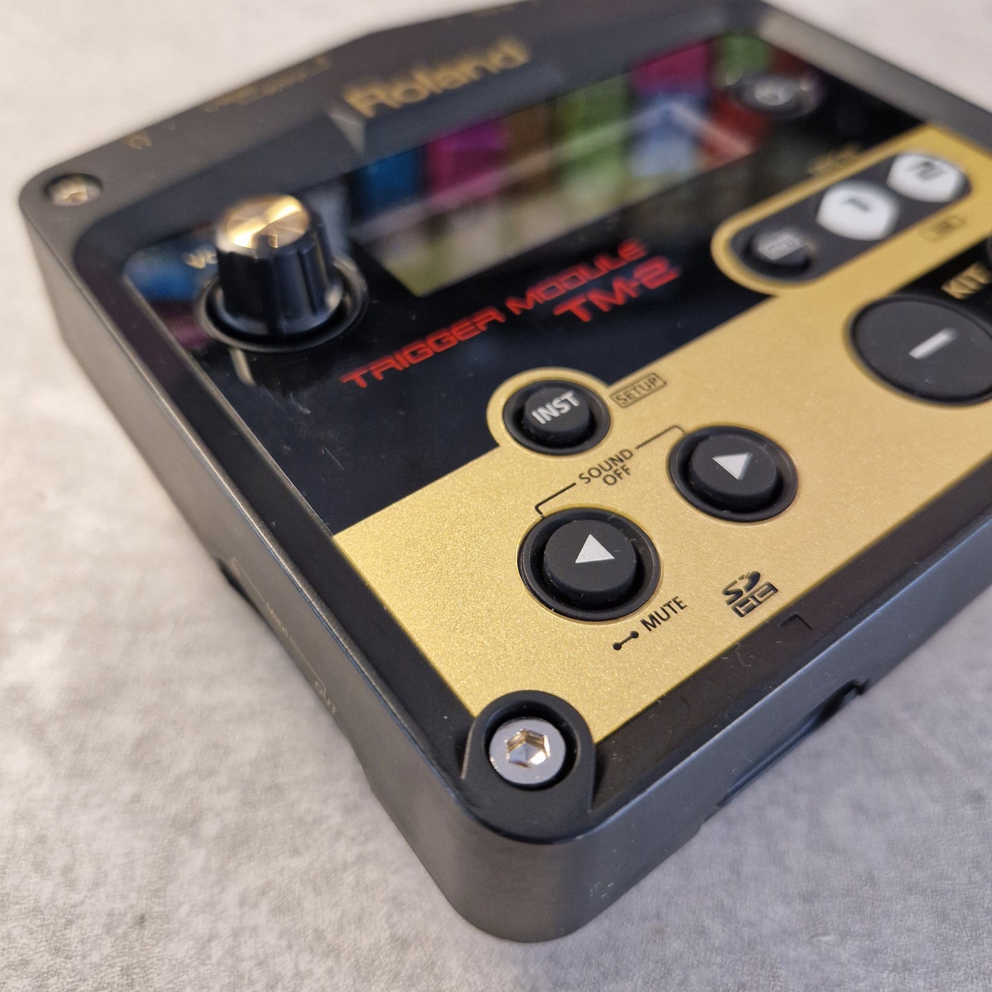 Pre-Owned Roland TM-2 A close-up of a Roland Music Man Time guitar modulation pedal, showing its gold and black faceplate with control buttons, a rotary knob, and a display screen, resting on a gray textured surface.