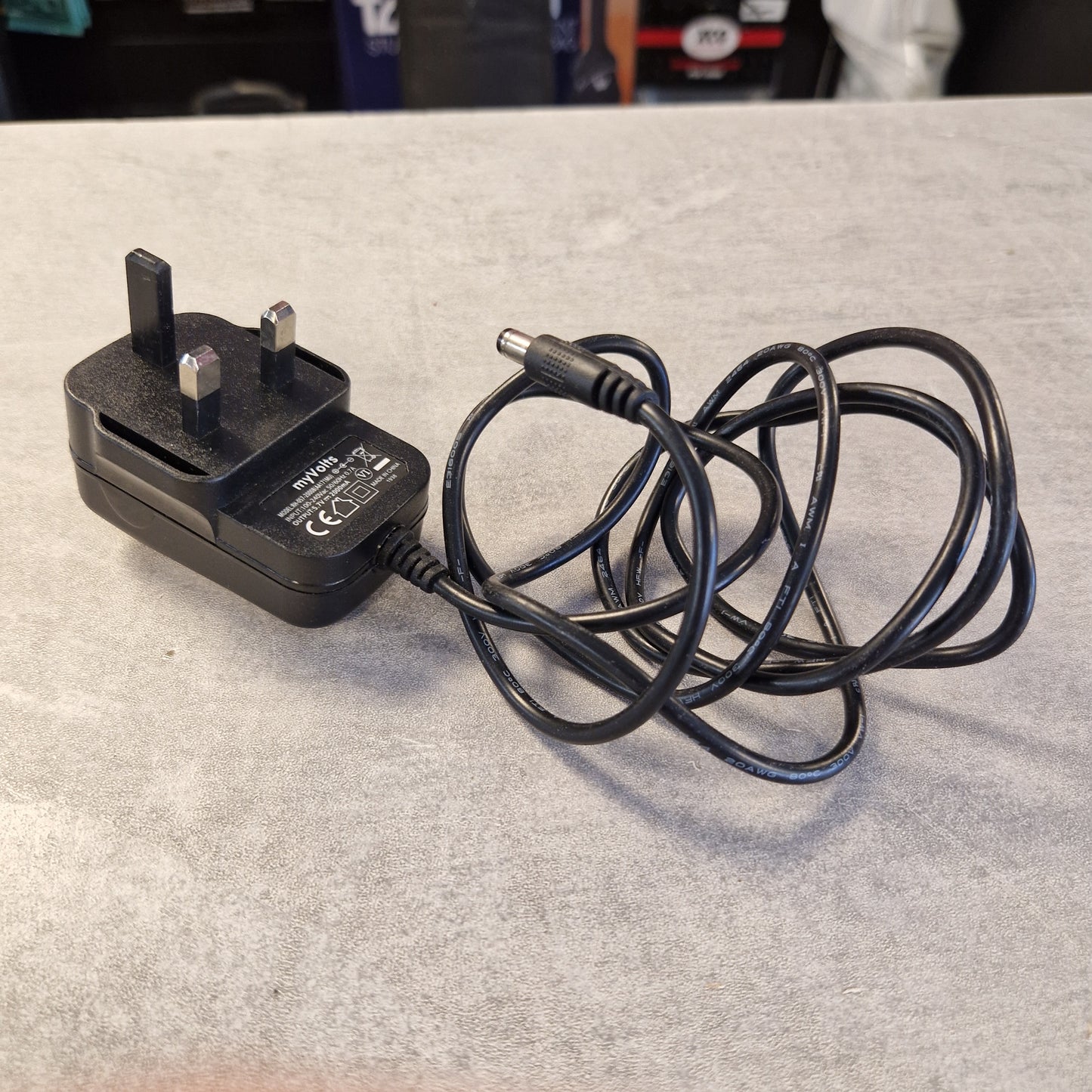 Pre-Owned Roland TM-2  A black UK three-pin wall charger adapter with a coiled power cord, lying on a gray textured surface.