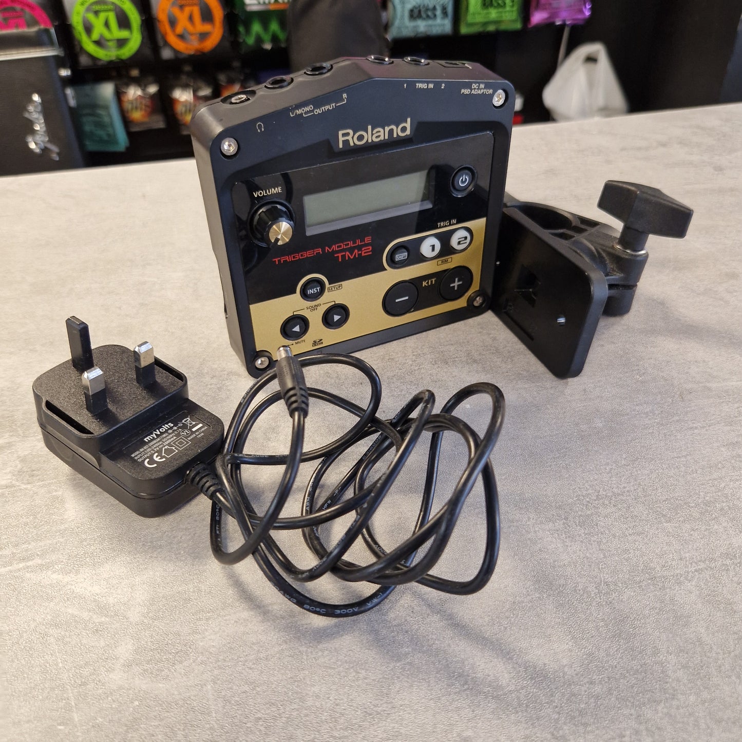 Pre-Owned Roland TM-2 Roland HP-5000A handheld multi-voice synthesizer in black with a gold control panel, shown with its black power cord and mounting clamp on a light gray surface.