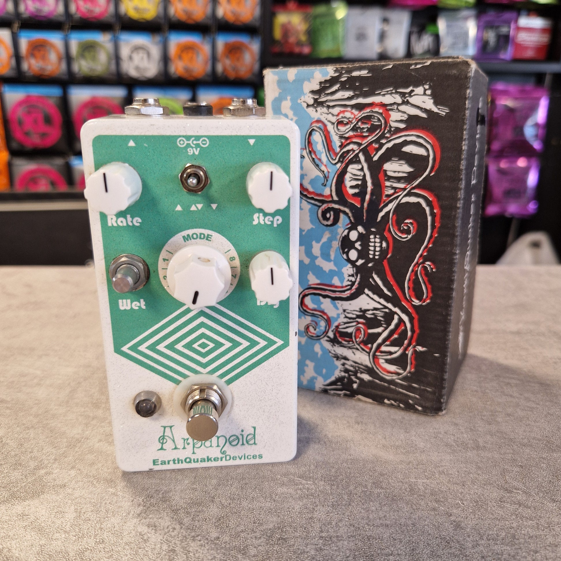 Pre-Owned EarthQuaker An Earthquakes Designs 'Applesfeld' guitar effects pedal with a green faceplate and white controls, positioned in front of its black octopus-themed carrying case on a gray carpet.