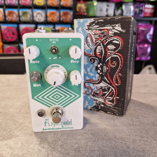 Pre-Owned EarthQuaker An Earthquakes Designs 'Applesfeld' guitar effects pedal with a green faceplate and white controls, positioned in front of its black octopus-themed carrying case on a gray carpet.