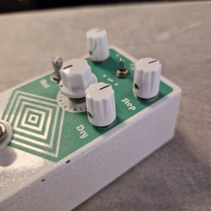 Pre-Owned EarthQuaker A close-up of a guitar effects pedal with a white metal casing, a green printed faceplate, and four white control knobs labeled Step, Dry, and others.