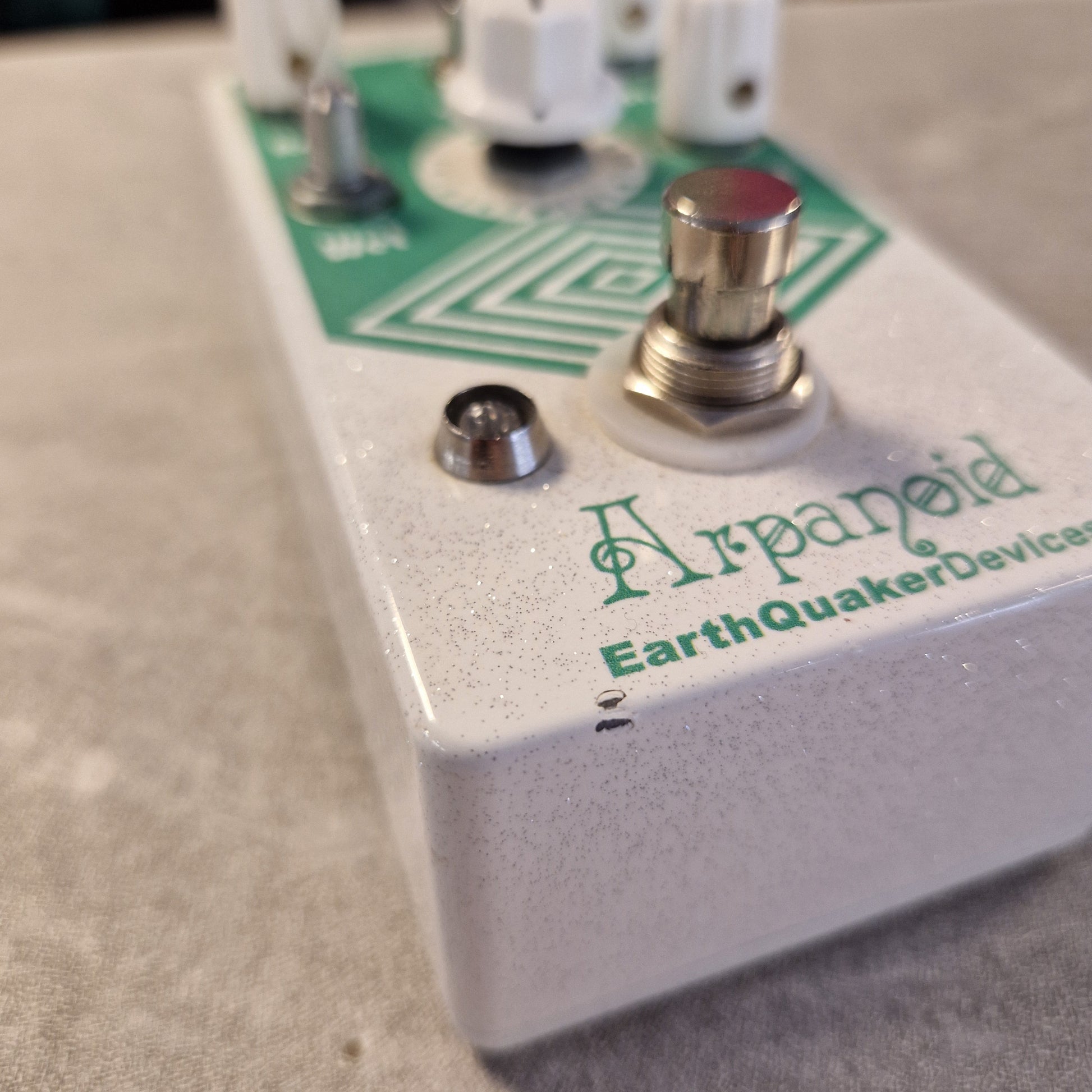 Pre-Owned EarthQuaker A close-up angled shot of an Earthquake Devices 'Japenoid' guitar effects pedal. The pedal has a light gray/speckled metal casing with green branding text and a green geometric logo, featuring a silver footswitch and a red-topped button.