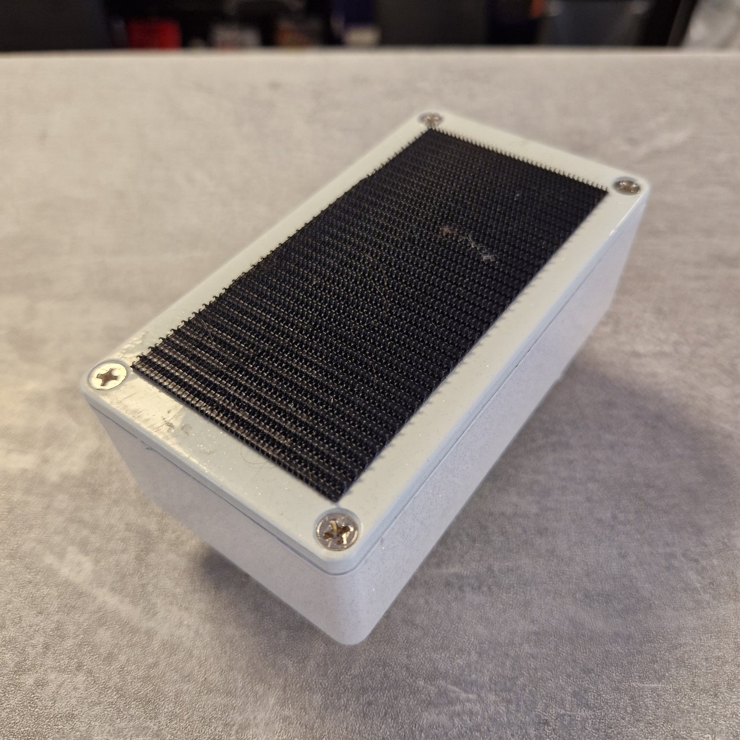 Pre-Owned EarthQuaker A rectangular black speaker grille mounted on a white metal box with four visible screws, photographed at an angle on a gray textured surface.