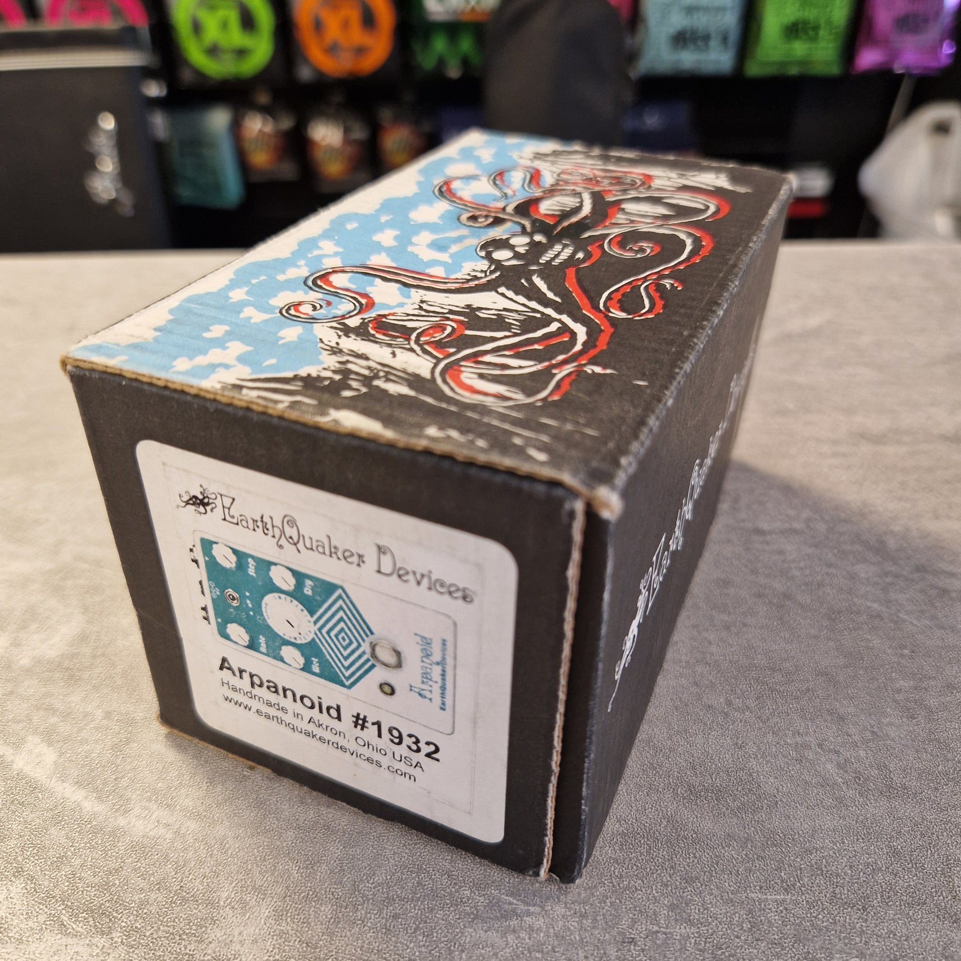 Pre-Owned EarthQuaker A product box for a Fuzz Face guitar pedal by Rusted Quiltz Devices, featuring a black cardboard base with red, white, and blue graphic artwork and a white product label showing the 'Ktranoid #1932' model.