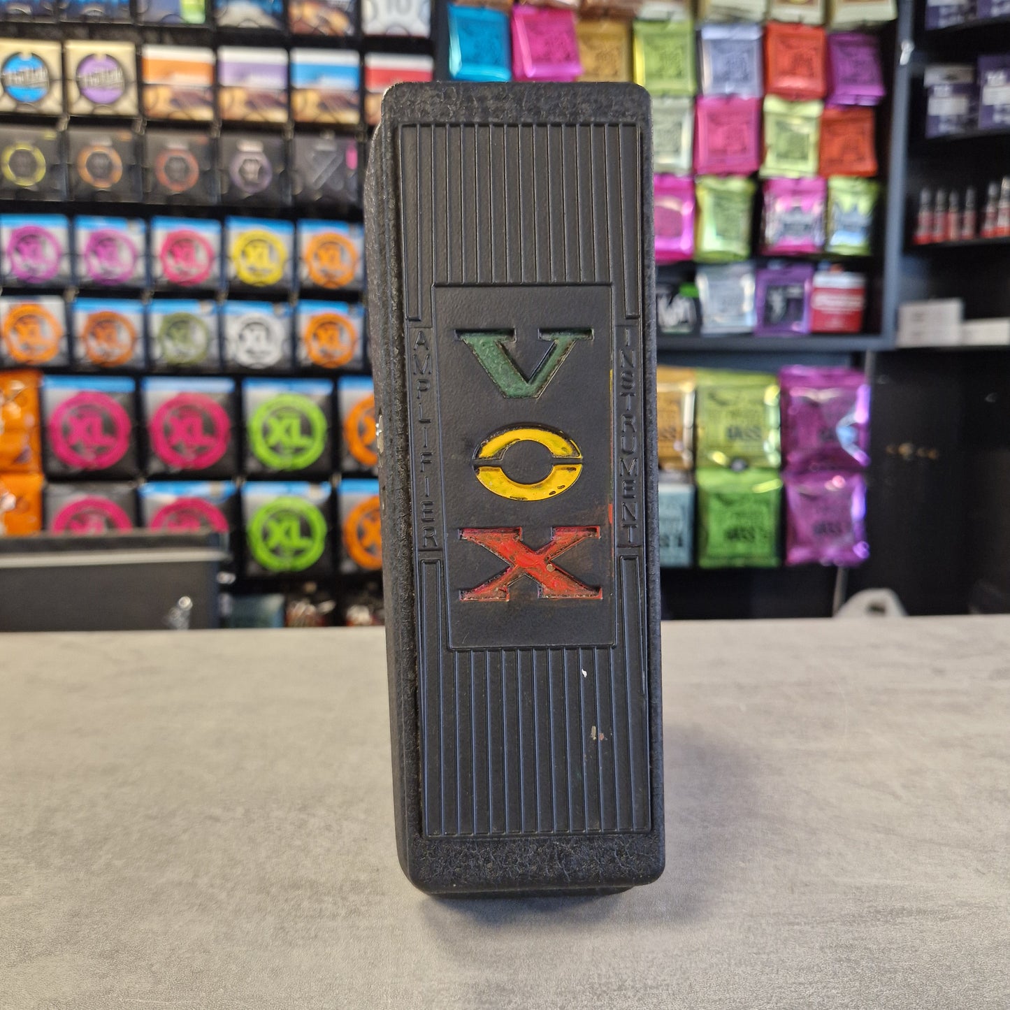 Pre-Owned Vox V845 A black rectangular vape pen with a textured ribbed body and a colorful 'DOX' logo on the front, shown in a retail store setting with colorful vape pods blurred in the background.