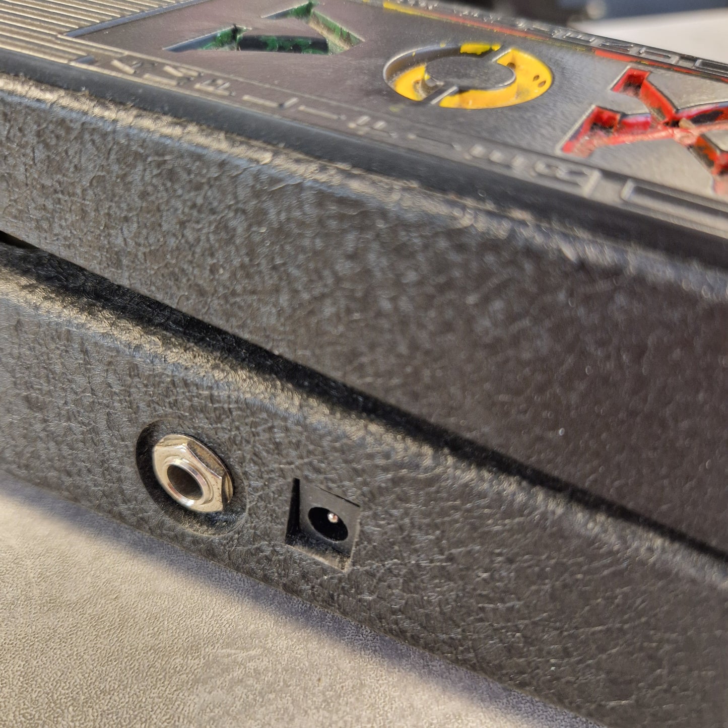 Pre-Owned Vox V845 Close-up of a textured dark gray or black panel with a small square black component and a round metal port, alongside part of a printed control panel with red, yellow, and green markings.