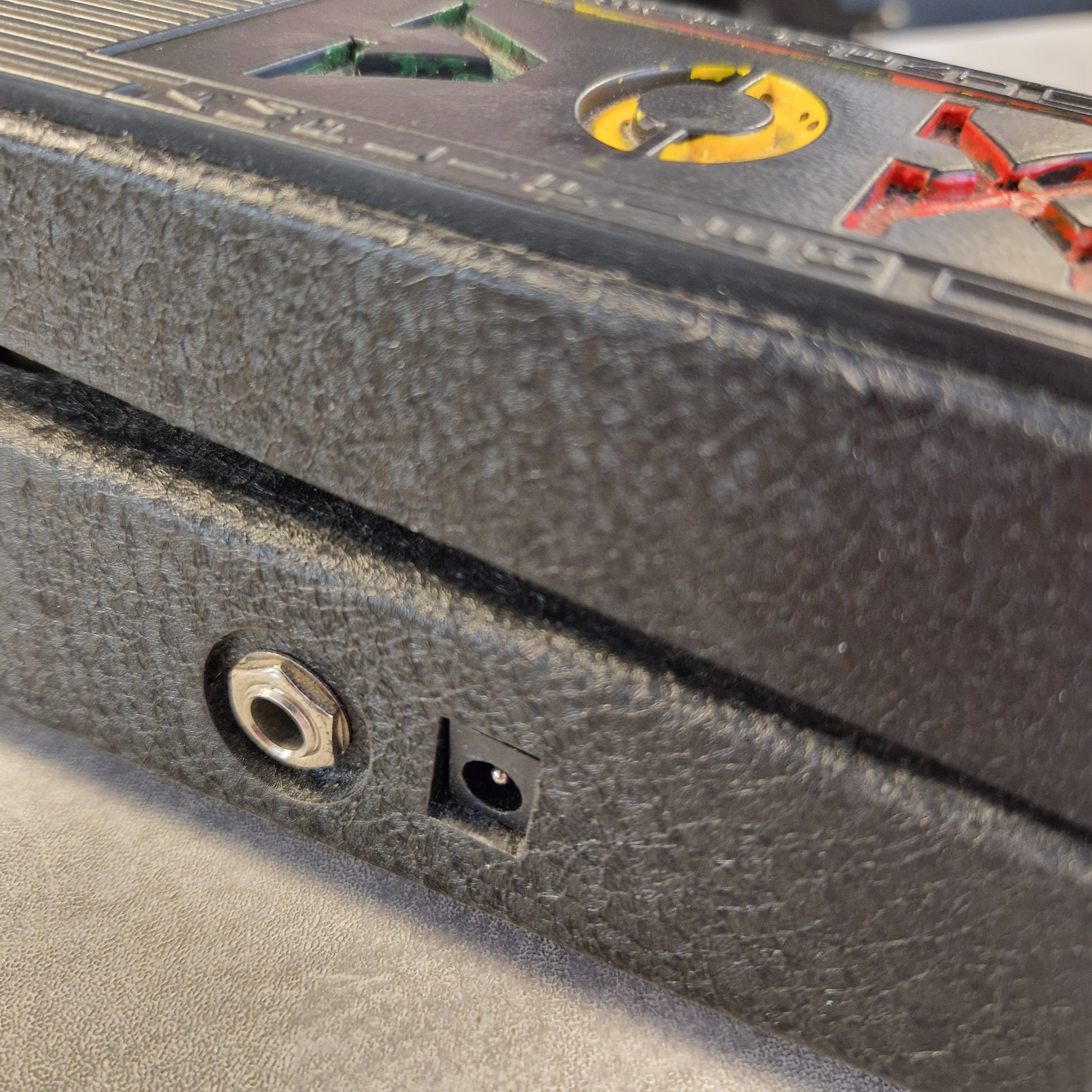 Pre-Owned Vox V845 Close-up of a textured dark gray or black panel with a small square black component and a round metal port, alongside part of a printed control panel with red, yellow, and green markings.
