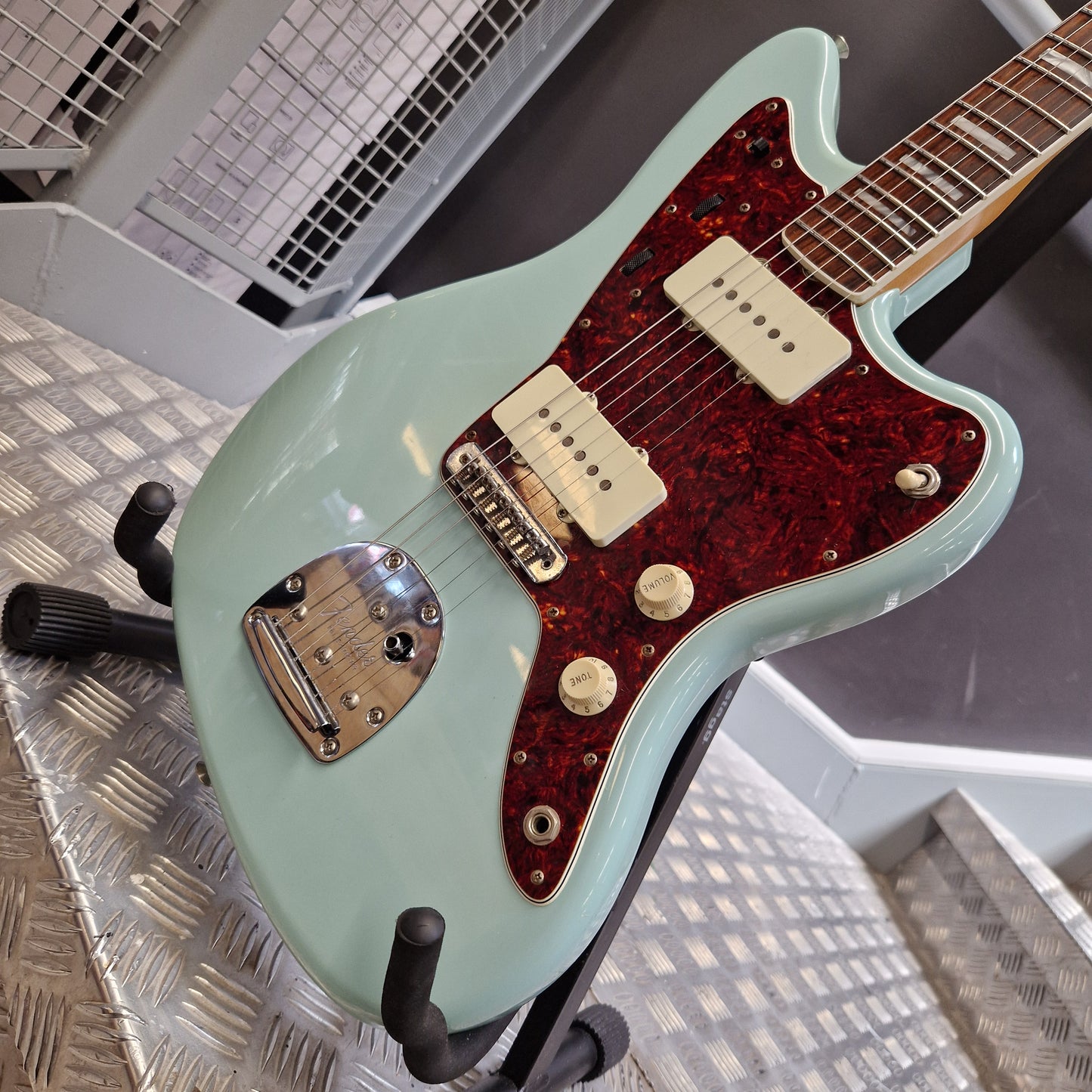 Pre-Owned Fender A mint green electric guitar with a red glitter pickguard, mounted on a stand on a diamond plate floor.