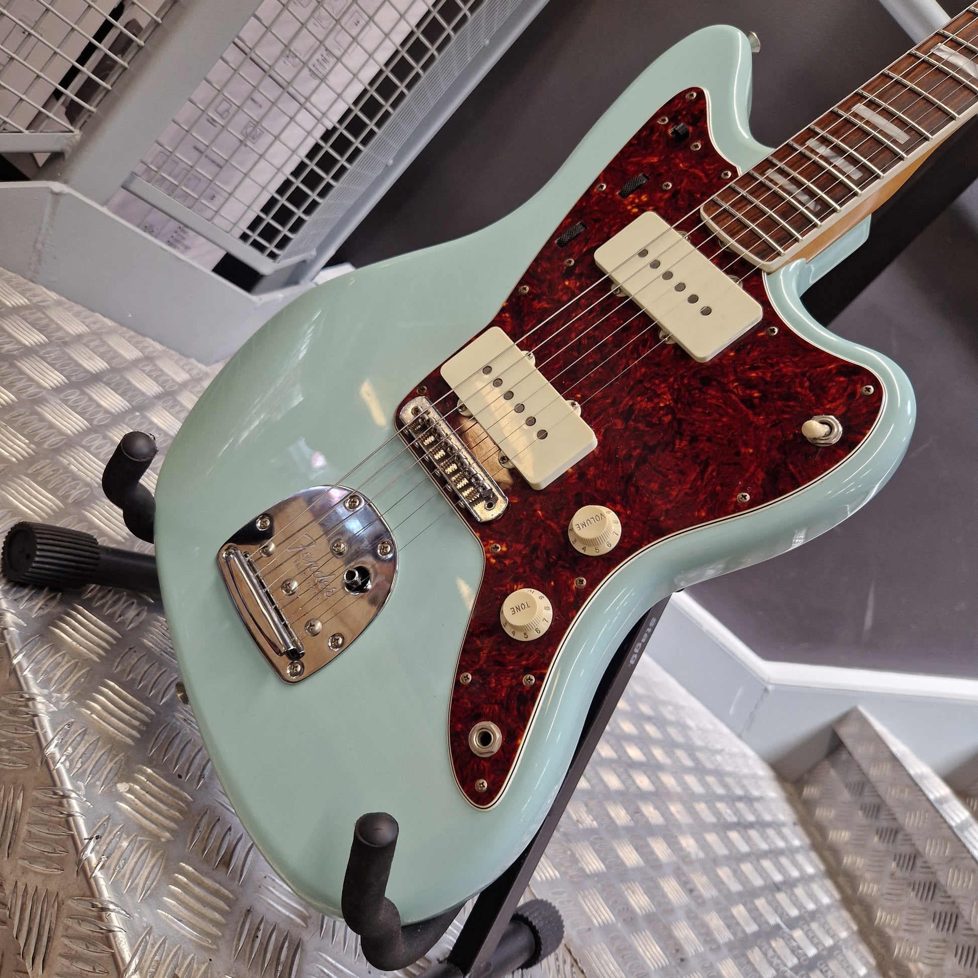 Pre-Owned Fender A mint green electric guitar with a red glitter pickguard, mounted on a stand on a diamond plate floor.